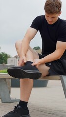 Wall Mural - Male athlete experiencing ankle pain after running, sitting on a bench outdoors and holding his injured leg, showing a sports sprain during workout, vertical high angle camera slow motion shot