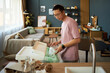 © DragonImages - Asian young adult man sorting packaging waste and placing plastic materials into recycling bin in modern living room, standing near kitchen counter with focused expression