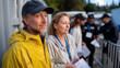 © Milos - A couple stands together in yellow and gray jackets, looking around at a bustling event scene filled with people and booths in the background, ready for a day out.