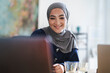 © Studio Marmellata - A happy young woman in a grey hijab smiles while working on her laptop. She is engaged in remote work or online learning from a bright home office. Empty glasses are on the desk.