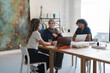 © Studio Marmellata - Three diverse young adults collaborate at a modern office table. Two women and one man work on laptops and discuss ideas in a bright, creative workspace.