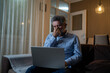 © Nenad - Stressed mature man sitting in living room at night with laptop