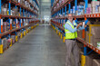 © wavebreak3 - Warehouse worker scanning boxes along aisle with tall blue and orange metal racks and yellow posts