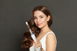 © artmim - Indoor portrait of woman with long wavy hairstyle using hair curling tongs iron, indoor portrait. Hairdresser equipment tools and hair styling concept