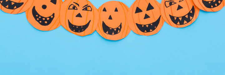 Wall Mural - Orange scary smiling pumpkin faces on light blue table background. Pastel color. Closeup. Creating Halloween decoration. Little child handcraft. Wide banner. Empty place for text. Top down view.