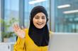 © Liubomir - Muslim businesswoman wearing a headset waving hand during an online video call, providing remote customer service or participating in virtual meeting from modern office