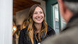 © dominic - Smiling woman greeting someone at the door with long hair wearing a black coat warmly in a well-lit indoor environment inviting atmosphere.