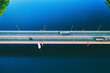 © nblxer - Aerial drone view of bridge asphalt road with cars and cargo trucks driving over blue river or lake water. Logistic and delivery concept.