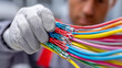 © Noteza - This image shows a close-up of an electrician wearing protective gloves and holding a bundle of colorful electrical wires. The wires, in shades of red, blue, yellow, green, orange, and gray, are neatl
