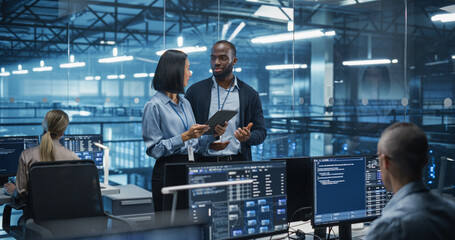 Male DevOps Engineer and Female IT Consultant Using a Tablet Computer, Standing in an Office Above the Rows of Servers in a Data Center, Developing Automation Scripts for Artificial Intelligence
