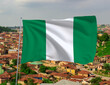 Nigerian flag waving on a pole above a city with rooftops and green surroundings, symbolizing patriotism, culture, and national pride in Nigeria’s urban and rural life