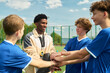 © Mediaphotos - Black man coaching group of teenage boys joining hands in team huddle on outdoor sports field, all wearing athletic uniforms and smiling, showing teamwork and encouragement