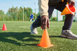 © Mediaphotos - Black man crouching on sports field placing orange training cone on artificial grass, holding additional cones in other hand, wearing athletic shoes, soccer players visible in background