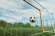 © Mediaphotos - Soccer ball hitting net inside outdoor goal on green field, capturing moment of scoring during game, metal goalposts and tall fence surrounding pitch under blue sky