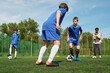 © Mediaphotos - Group of teenage boys playing soccer on outdoor field, one dribbling ball while others defending and observing, Black man standing on sideline watching training session