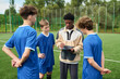 © Mediaphotos - Black man coaching three teenage boys on outdoor soccer field, holding clipboard and explaining strategy while teenagers listening attentively during practice