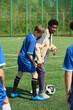© Mediaphotos - Black man coaching boy during soccer practice on outdoor field, demonstrating footwork with soccer ball while other children observing training session