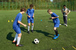 © Mediaphotos - Three teenage boys practicing soccer dribbling drills on outdoor field while Black male coach observing training session, green fence and trees in background