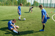© Mediaphotos - Group of teenage boys playing soccer on outdoor field, one teenager sitting on grass stretching leg while others standing nearby, soccer ball and cones visible in background