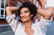 © BullRun - Smiling woman in white shirt holding hair with both hands, looking up with joy. Conceptual stock image reflecting authenticity, individuality, optimism, empowerment, and modern urban lifestyle.