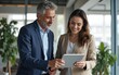 © David - Team leader explain trainee working process using tablet pc computer. Two professional business people working together. Partners mature man, woman discussing project in office workplace. Copy space