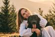 © KatyaPulka - Happy young woman hugging her black Labrador dog outside in nature. Smiling girl and her dog enjoying a beautiful sunny day with trees and mountains in the background.