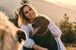 © KatyaPulka - A smiling young woman embraces her happy black Labrador Retriever in a scenic outdoor setting with mountains and trees. A second dog is partially visible. Golden light enhances the warm feeling.
