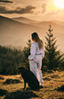© KatyaPulka - A woman in a sweatsuit standing with her black dog in a mountain landscape at sunset. Golden hour light illuminates the beautiful view. Peaceful, serene moment.