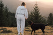 © KatyaPulka - Woman and her dog enjoying the mountain views at sunrise. Back view of a girl in a grey tracksuit and a black labrador on a hill. Scenic landscape and freedom concept.