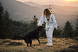 © KatyaPulka - A woman plays fetch with her black labrador dog on a mountain top at sunset. Hiking and outdoor activities concept, enjoying nature and companionship with a pet in scenic landscape.