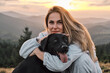 © KatyaPulka - A heartwarming moment of a young woman embracing her black Labrador Retriever against a stunning mountain sunset. Pet love, outdoor adventure, and companionship.