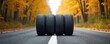 © Raisa - Four car tires stacked on asphalt road surrounded by autumn forest. Vibrant yellow, orange leaves line roadside, with trees forming colorful tunnel. Scene suggests tire inspection seasonal switch,