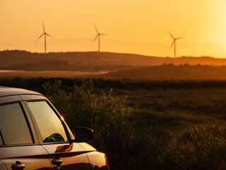 Wall Mural - A red car is parked in a field with a sunset in the background. The car is facing the sun, and the sky is orange and pink. Wind farm turbines in the background. Sightseeing modern technology concept