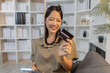 © kamiphotos - A cheerful young woman using her smartphone for online shopping, holding a credit card while smiling with excitement, sitting comfortably indoors and enjoying digital convenience.