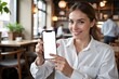© YiXiong - A woman in a white shirt holding a blank smartphone in a cozy cafe.