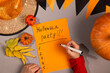 © olga_sova - A man writes a list on orange paper with the inscription - Halloween Party next to a pumpkin, miniature pumpkins, autumn leaves and a witch's hat.