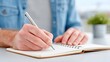 © Xion - Close Up of Male Hand Writing in Open Notebook with Pen on White Table and Blurred Green Plant Background in Natural Lighting