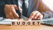 © SnapGlint - Hands adjust wooden “BUDGET” blocks over dollar bills—scissors poised to cut between “D” and “G” evoke financial reduction, expense control, and strategic adjustment in minimalist economic symbolism c