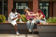 © DragonImages - Group of three teenager students, Black male and two Caucasian friends, sitting outdoors on bench, laughing and talking while young woman holding drink and gesturing toward friends