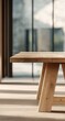 © adha - Close-up of a light-brown wooden table with a simple, wide rectangular top and wide, angled legs. Blurred view of a room with windows and out-of-focus natural scenery