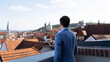 © VladyslavShcherbakov - Man in blue jacket standing on terrace overlooking historic city with red rooftops and cathedral domes, symbolizing travel inspiration, culture, and urban exploration.