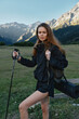 © SHOTPRIME STUDIO - A young woman stands in a mountainous meadow, holding a trekking pole with a rolled mat on her back. She wears a black jacket and shorts, looking ahead with confidence for an outdoor adventure.