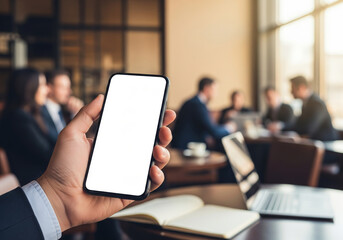 Ground level perspective of a hand holding smartphone with empty white screen, office team meeting blurred in background with laptop and notebook on table, business app mockup concept