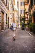 © StockMediaSeller - Woman with backpack and straw hat walking along a narrow cobblestone street in Rome surrounded by plants and historic houses