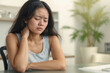 © polack - Asian woman with neck pain is sitting at a table at home. She looks down and it hurts