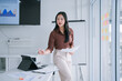 © MINAE - Asian businesswoman standing in a modern office, holding documents and a pen, presenting financial data and charts displayed on a screen during a corporate meeting