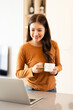 © Fahng - Young woman drinking morning coffee and looking at monitor of laptop while sitting in the kitchen at home.
