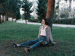 © SHOTPRIME STUDIO - A thoughtful woman sits beneath a tree in a quiet park, dressed in casual attire. She rests on the grass, gazing ahead, conveying calmness and reflection in a natural outdoor setting.