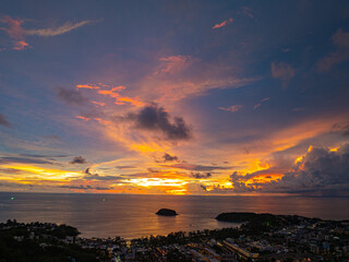 Wall Mural - A breathtaking sunset paints the sky in vivid hues of orange and blue over a coastal town, with lush green hills framing the scene. A distant hilltop structure adds a serene tranquil landscape.