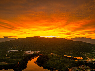 Wall Mural - A breathtaking aerial view of a vivid orange sunset casting dramatic light over forested hills and a reflective river, with urban elements nestled at the landscape’s edge beneath a glowing sky.